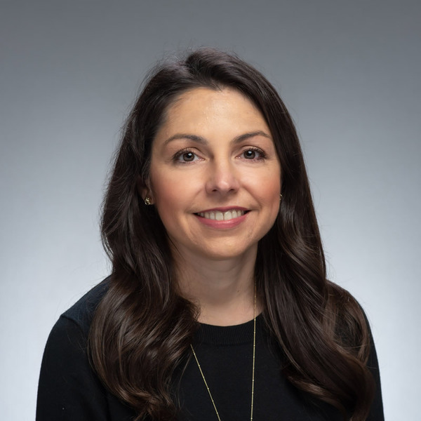 Jill Pentimonti. A smiling woman with long, wavy dark brown hair and gold earrings wears a black sweater and delicate gold necklace against a gray background.