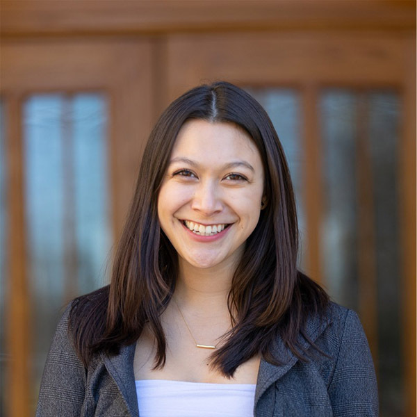 Sophia Alvarez. A young woman with dark hair, wearing a white top and gray plaid blazer, smiles broadly against a blurred wooden door background.